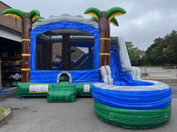 A large inflatable bounce house with a blue and gray slide, decorated with palm trees, is set up outdoors on pavement near a building and greenery.