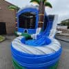 An inflatable water slide with a tropical theme, featuring palm trees and a curved blue and gray slide ending in a small pool, set up outdoors on pavement near a brick building.