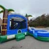 An inflatable bounce house and slide with a blue and green tropical theme, featuring palm trees, set up outdoors on a gray pavement near a building and trees under a cloudy sky.