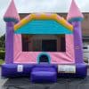A colorful inflatable bounce house shaped like a castle, with pink, purple, and blue walls and two turret towers, set up outdoors on a black mat near a brick building.