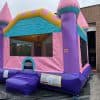 A large, colorful inflatable bounce house designed like a castle with pink turrets, a yellow and blue roof, and purple walls, set up outdoors on a black mat near an industrial building.