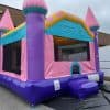 A colorful inflatable bounce house with pink towers, purple and blue walls, and a small entrance sits on pavement outside a brick building under a cloudy sky.