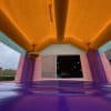 View from inside a colorful inflatable bounce house with purple floor and yellow-orange ceiling, looking out at a warehouse building and some greenery outside through the open entrance.