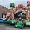 An inflatable bounce house styled like a Minecraft structure, featuring large pixelated heads of a Creeper and a pig, with a small slide attached, set up outdoors near a warehouse.