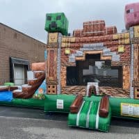 An inflatable bounce house styled like a Minecraft structure, featuring large pixelated heads of a Creeper and a pig, with a small slide attached, set up outdoors near a warehouse.