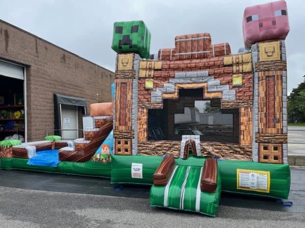 An inflatable bounce house styled like a Minecraft structure, featuring large pixelated heads of a Creeper and a pig, with a small slide attached, set up outdoors near a warehouse.