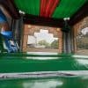 A photo taken from inside an inflatable bounce house with shiny green and blue surfaces, netted windows, and a view of trees and buildings outside through a castle-themed opening.