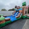 A large inflatable slide outdoors decorated with pixelated, Minecraft-inspired graphics, featuring blocky characters and a blue sliding area. The slide is set up on a black tarp next to buildings and trees.