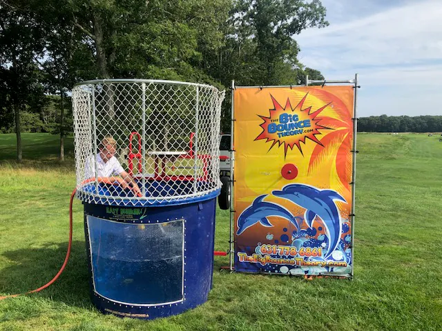 A person sits inside a dunk tank filled with water, surrounded by a metal cage, on a grassy field in NY. The tank features a colorful sign with dolphins and the text "The Big Bounce Theory.com," perfect for party rentals Long Island style. Trees are visible in the background.