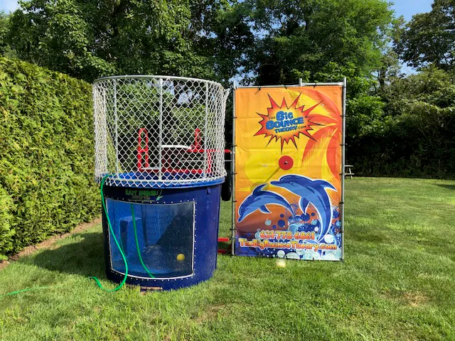 A dunk tank with a blue base and white metal cage stands on grass next to a colorful sign featuring dolphins and the words “Big Bounce Thunder”—perfect for party rentals Long Island. A garden hose lies in front, with trees and bushes in the background.