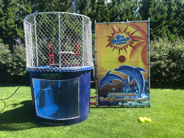 A dunk tank with a blue base and metal cage is set up on grass. A colorful dolphin-themed backdrop with a phone number stands behind it. Three yellow balls lie in front—perfect for party rentals Long Island or any fun NY event!.