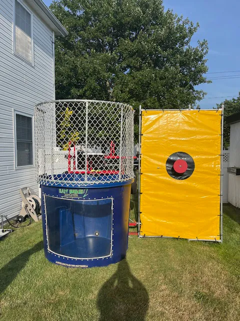 A blue dunk tank with a transparent window and metal cage is set up on grass next to a large yellow target backdrop in a backyard, perfect for party rentals Long Island, with trees and houses in the background.
