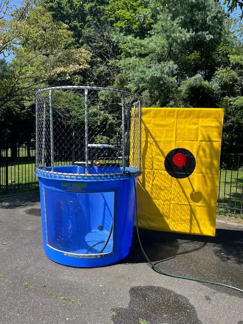 A blue dunk tank from party rentals Long Island, NY sits outdoors beside a bright yellow bullseye target board and a green hose. Trees and a black fence form the backdrop, perfect for your next event with bounce house Long Island fun.