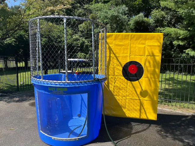 A blue dunk tank with a metal cage sits beside a large yellow target board outdoors on a sunny day, perfect for party rentals Long Island, NY, with trees and a black fence in the background.