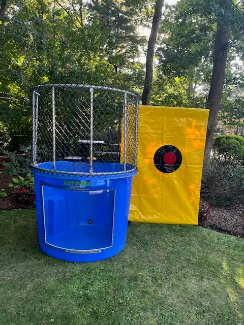 A blue dunk tank with an open window and a metal cage sits on grass beside a yellow target backdrop in a wooded outdoor area, perfect for NY parties or paired with bounce house Long Island rentals.