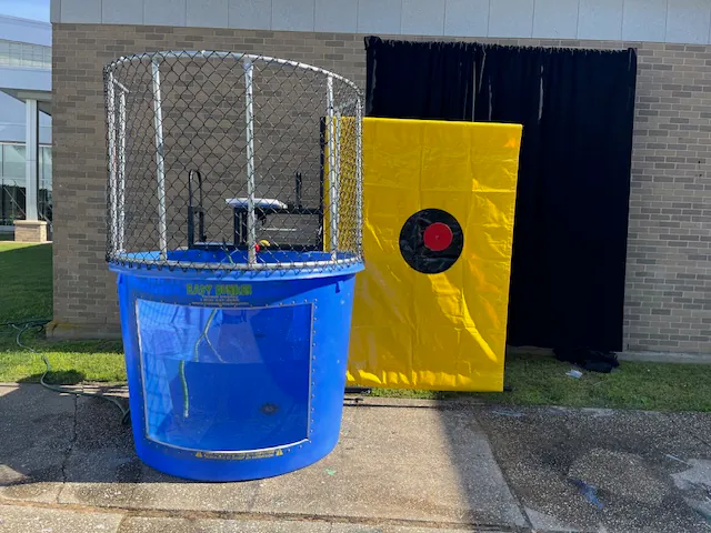 A blue dunk tank with a metal safety cage is set up outdoors. Next to it is a yellow target panel with a red and black bullseye—perfect for party rentals Long Island events—in front of a black curtain and brick wall.