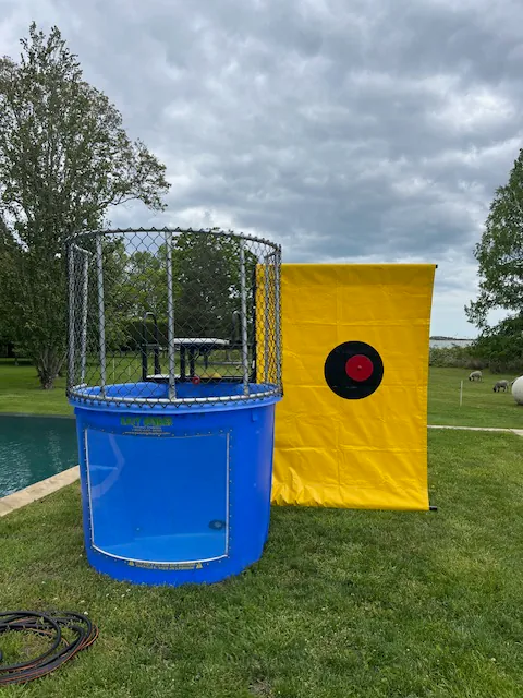 A blue dunk tank with a metal cage stands on grass beside a large yellow target backdrop, perfect for party rentals Long Island, NY. Set outdoors near a pool and surrounded by trees under a cloudy sky.