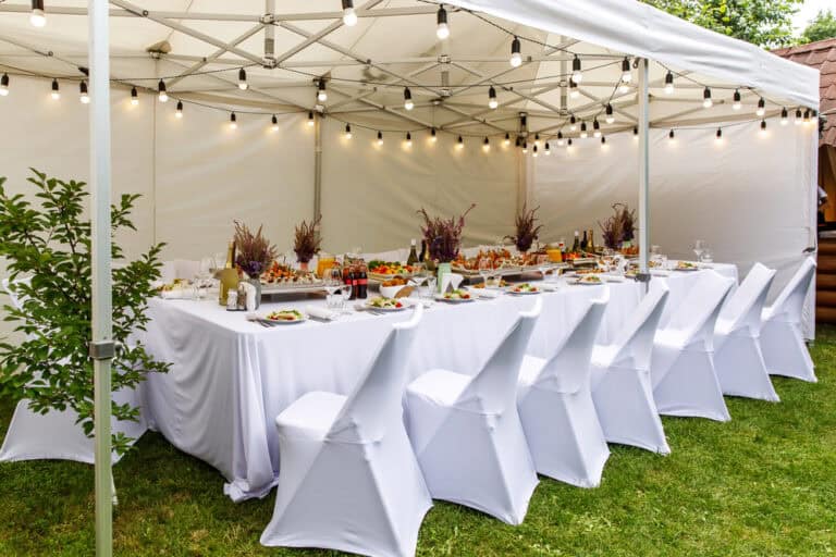 A long table covered with a white tablecloth is set for a meal under a white canopy tent, with white covered chairs and string lights overhead. Perfect for celebrations, party rentals Long Island can enhance your event with decor, food, and seating in NY style.