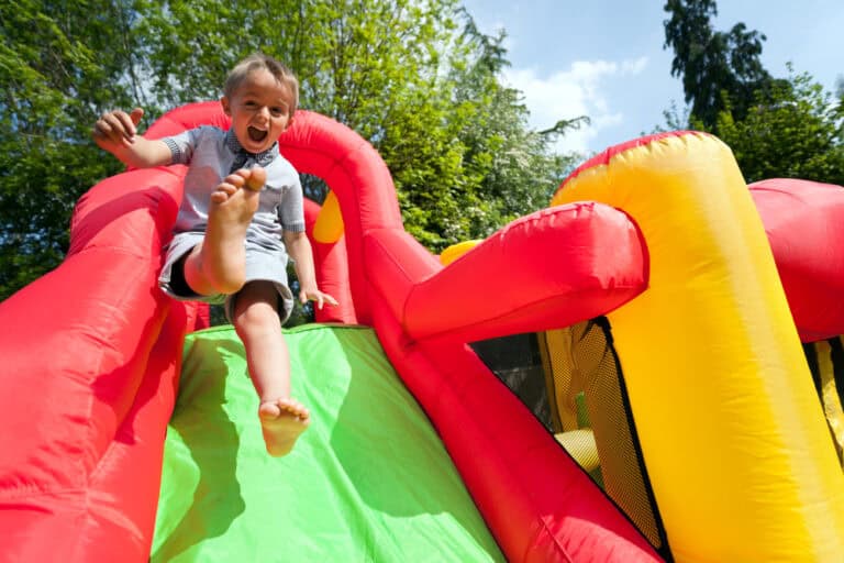 A young child joyfully slides down a green inflatable slide, part of a bright red and yellow bounce house Long Island, outdoors on a sunny NY day with trees and blue sky in the background.