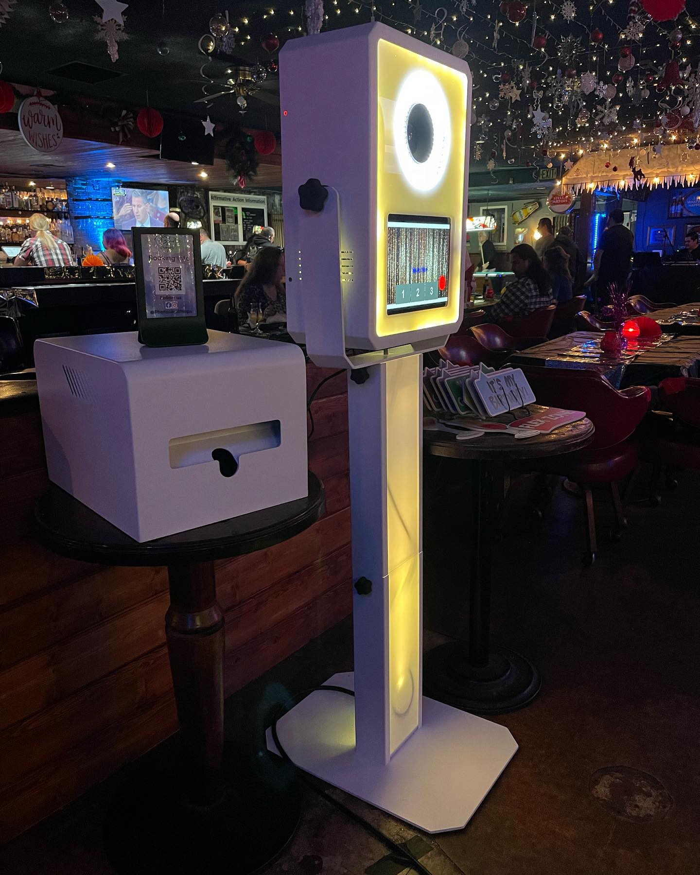 A modern photo booth with a glowing ring light stands next to a white printer on a small table in a festive bar decorated with holiday ornaments and lights. People are seated and socializing in the background.