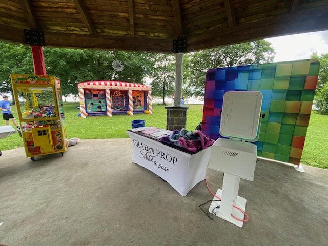 A photo booth setup under a gazebo features a table with props, a white kiosk, a checkered backdrop, and a colorful bounce house and claw machine on grass in the background. Trees and a lake are visible beyond the booth.