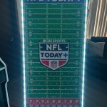 A large, vertical NFL Today+ sign with a football field design, lighted border, and a CBS Sports logo, standing indoors near a window and some furniture.