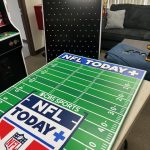 A game board resembling a football field labeled "CBS Sports NFL Today+" sits on a table in an office, with a couch, arcade machine, and pegboard visible in the background.