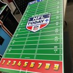 A game board designed like an American football field with CBS Sports NFL Today+ branding, yard lines, and numbers 1 to 9 along a red strip at the bottom, placed on a table indoors.