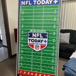 A large NFL-themed Plinko board labeled "CBS Sports NFL Today+" stands in a room, featuring a football field design, metal pegs, and numbered slots from 1 to 9 at the bottom. A couch and window blinds are in the background.
