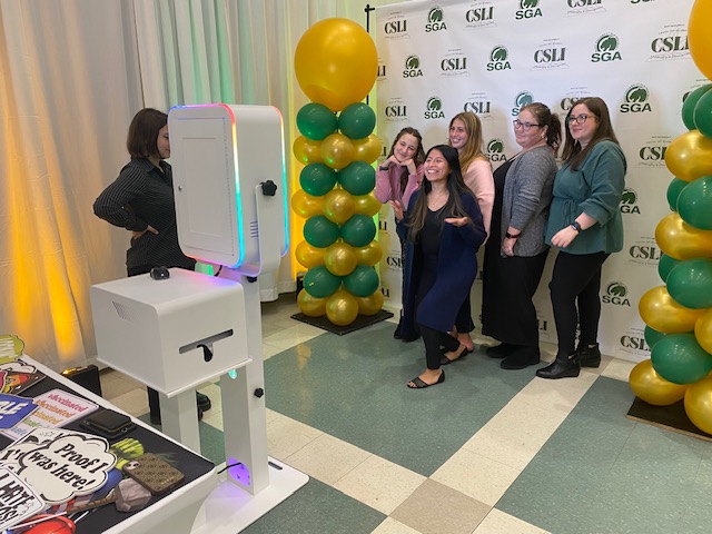 Six people pose and smile for a photo at a photo booth with colorful lights. Yellow and green balloons and a backdrop with “CSLI” and “SGA” logos are in the background. Photo props are arranged on a nearby table.