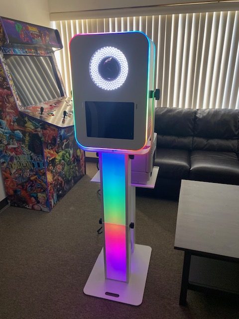 A colorful photo booth with a ring light stands in a room. An arcade game is in the corner, and a black couch sits nearby in front of vertical window blinds. The photo booth glows with rainbow LED lights.