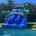 A large inflatable water slide with blue and white colors and palm tree decorations stands next to a pool. A child in a swimsuit walks nearby on the sunny, grassy yard with trees in the background.