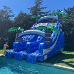 A large blue inflatable water slide with palm tree decorations stands next to a swimming pool. One person is sliding down, while another stands at the pool's edge on a sunny day. Trees surround the backyard.