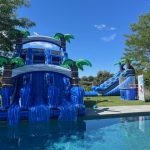 Two large inflatable water slides with palm tree decorations are set up beside a swimming pool on a sunny day, surrounded by green grass and trees. The sky is clear and blue.