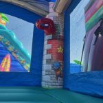 Inside a blue inflatable bounce house, there's a pillar decorated with pixel art of a character climbing bricks and various icons. Through the mesh window, an inflatable slide with beach and surf designs is visible outside.