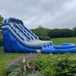 A large inflatable blue and gray water slide is set up on a grassy lawn, with colorful flowers in the foreground and trees and bushes in the background under a cloudy sky.