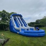 A large blue and white inflatable water slide is set up on a green lawn, with a small pool at the bottom. Two people are near the slide, and trees and plants border the grassy area under a cloudy sky.