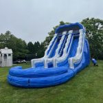 A large inflatable blue and white water slide is set up on a grassy lawn, with trees and a playhouse visible in the background. A person is standing on the right side near the slide.