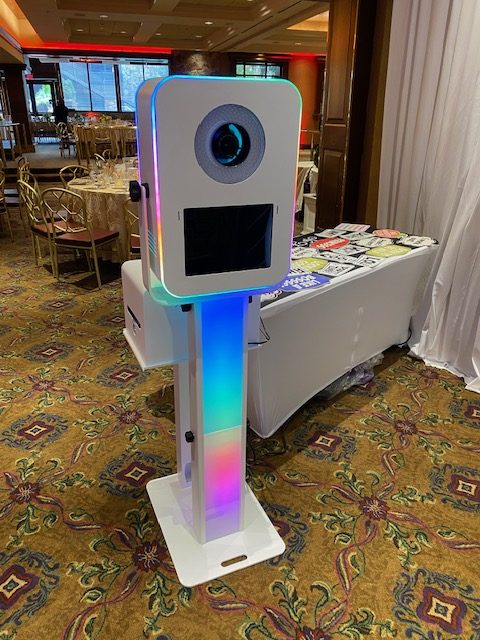 A colorful LED photo booth with a camera and screen stands on a decorative carpet at an indoor event venue with tables, chairs, and a table of props in the background.