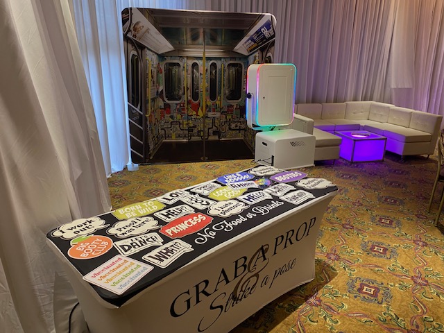 A photo booth setup at an event with a subway-themed backdrop, a table of playful photo props, and white couches around glowing cube tables on a patterned carpet, all surrounded by white curtains.