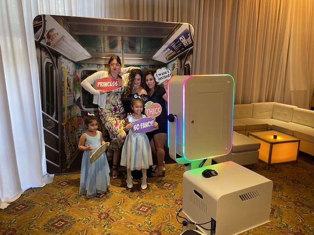 Four people and two children pose in front of a subway-themed photo booth backdrop, holding colorful signs with phrases like "PRINCESS" and "SO FANCY!" The scene is festive, with a sofa and glowing table nearby.