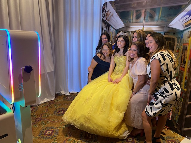 A young woman in a yellow ball gown and crown poses with five smiling women in front of a photo booth, against a backdrop of subway murals and draped white curtains.