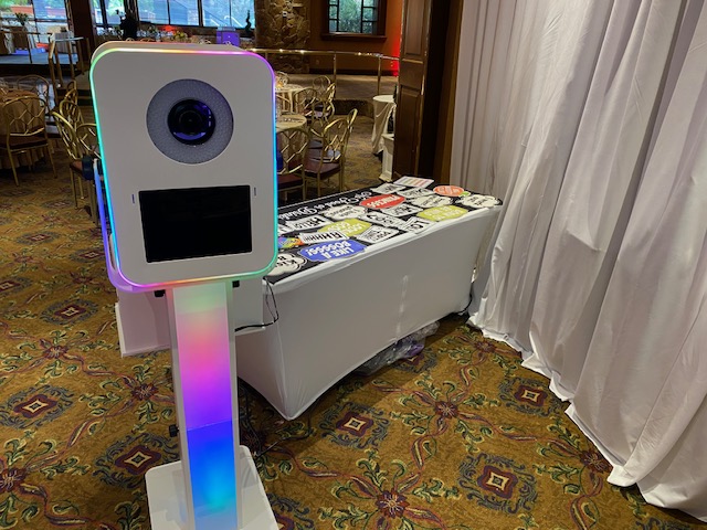 A colorful photo booth with lights stands next to a table covered with various photo props and signs at an indoor event venue with patterned carpet and gold chairs.