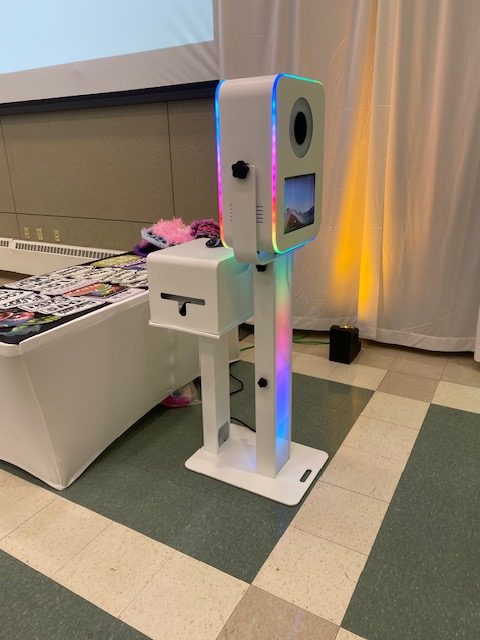 A photo booth with colorful LED lights stands next to a table with props in a room with tiles and a curtain backdrop. A small screen and camera lens are visible on the booth's front.