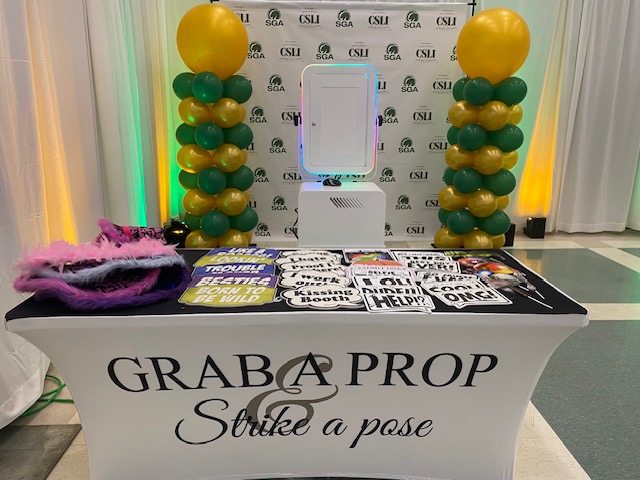 A photo booth setup with a table labeled "GRAB A PROP, Strike a Pose." The table holds colorful props and signs, with green and yellow balloon columns and a branded backdrop in the background.