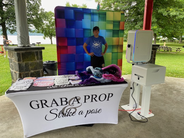 A man stands smiling in front of a colorful photo booth backdrop in a park pavilion. A table with props and a sign reading “Grab a Prop & Strike a Pose” is set up next to a photo booth machine. Trees and picnic tables are in the background.