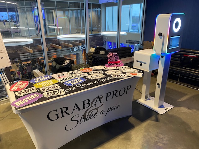 A photo booth setup at an event with a table displaying various colorful photo props, a sign reading "GRAB A PROP Strike a pose," and a modern photo booth machine with a touchscreen and ring light.