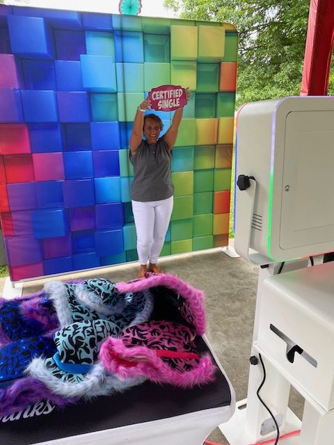 A woman stands in front of a colorful, tiled backdrop, smiling and holding up a sign that says "CERTIFIED SINGLE." Photo props with animal print and fur are on a nearby table, and a photo booth setup is visible.
