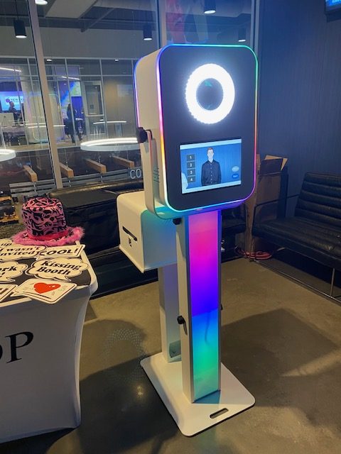 A colorful photo booth with LED lights, a ring light, and a screen displaying a person’s image stands indoors next to a table with props and stickers.