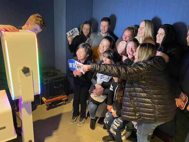 A group of people pose closely together in front of a photo booth, smiling and holding props with text. A bright light from the booth illuminates their faces in a dimly lit room with blue walls.